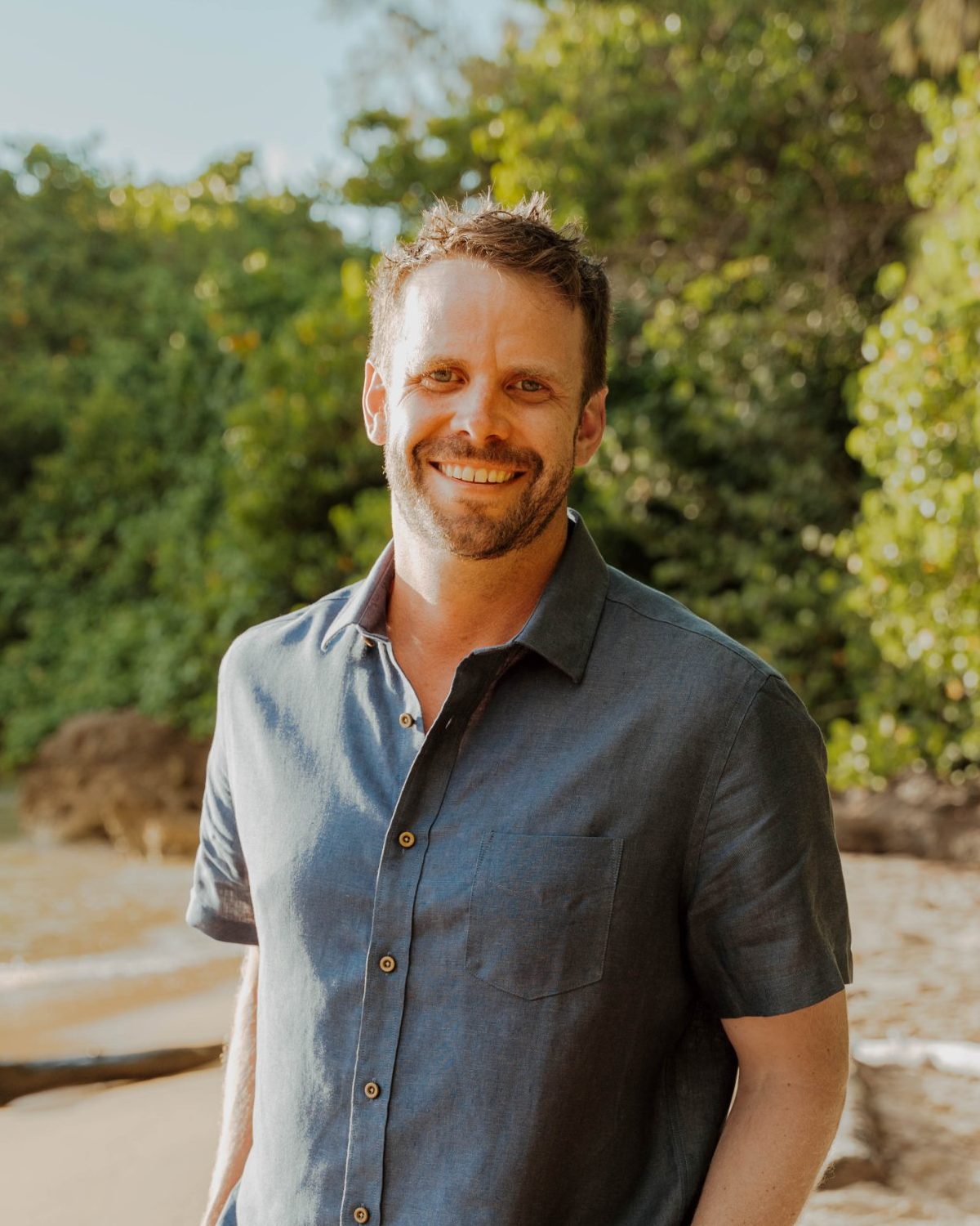 Max Cowan on a beach, smiling, in a blue linen shirt